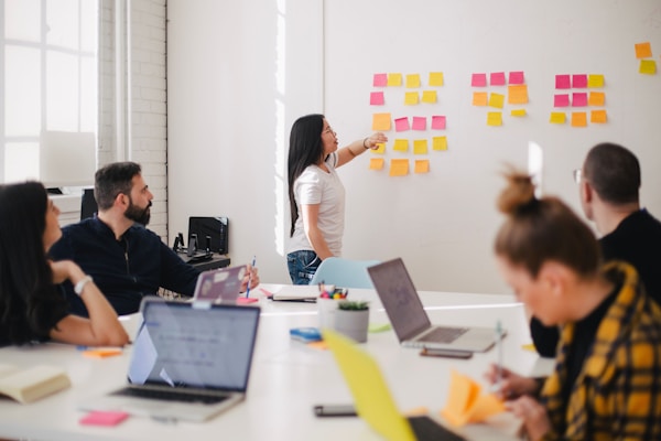 Team in a bright office discussing ideas as a woman points to a wall covered with sticky notes on a whiteboard area.