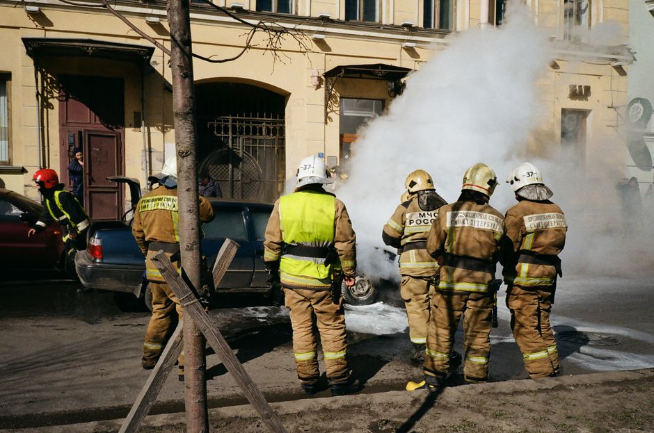Firefighters in full turnout gear fight a car fire on a city street, thick smoke pouring from the vehicle, nearby buildings in view.