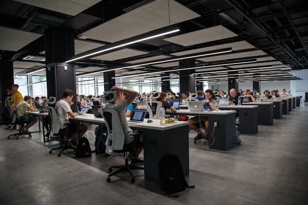 Open-plan office with rows of desks and people working on laptops under modern ceiling lights.